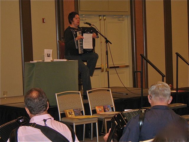 Debra Peters presenting her “Accordion Bass Patterns” DVD during a workshop at the National Accordion Association (NAA) convention, 2010. She is seated on stage playing accordion with sheet music and microphone.